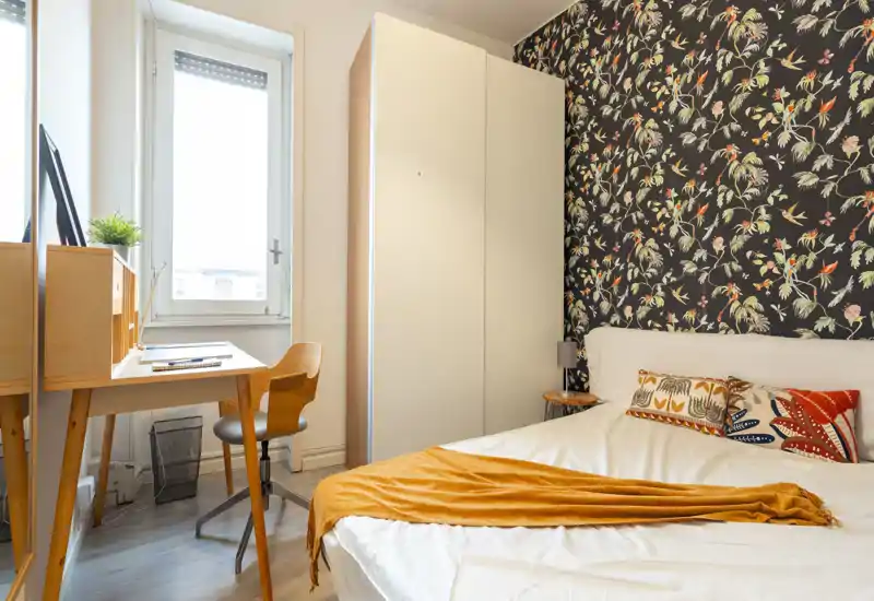 Bright, stylish bedroom with a comfortable double bed, patterned accent wall and a compact wooden desk by the window — ideal for working and resting.