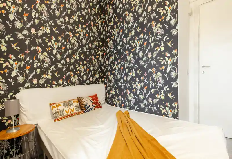 Bright, stylish bedroom with patterned accent wallpaper, a comfortable double bed dressed with decorative cushions and a warm throw.