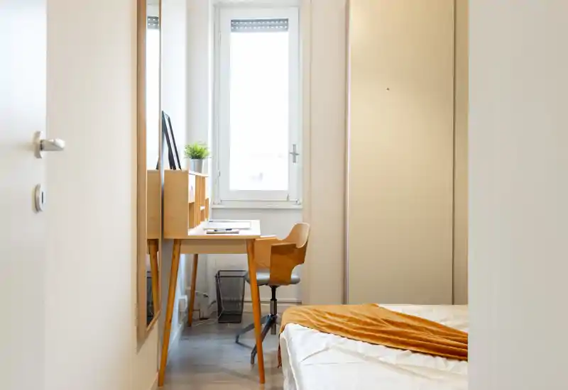 Bright, minimal bedroom with a wooden desk by the window and a neatly made bed with a warm throw. The space features clean lines, natural light and simple Scandinavian-inspired furniture.