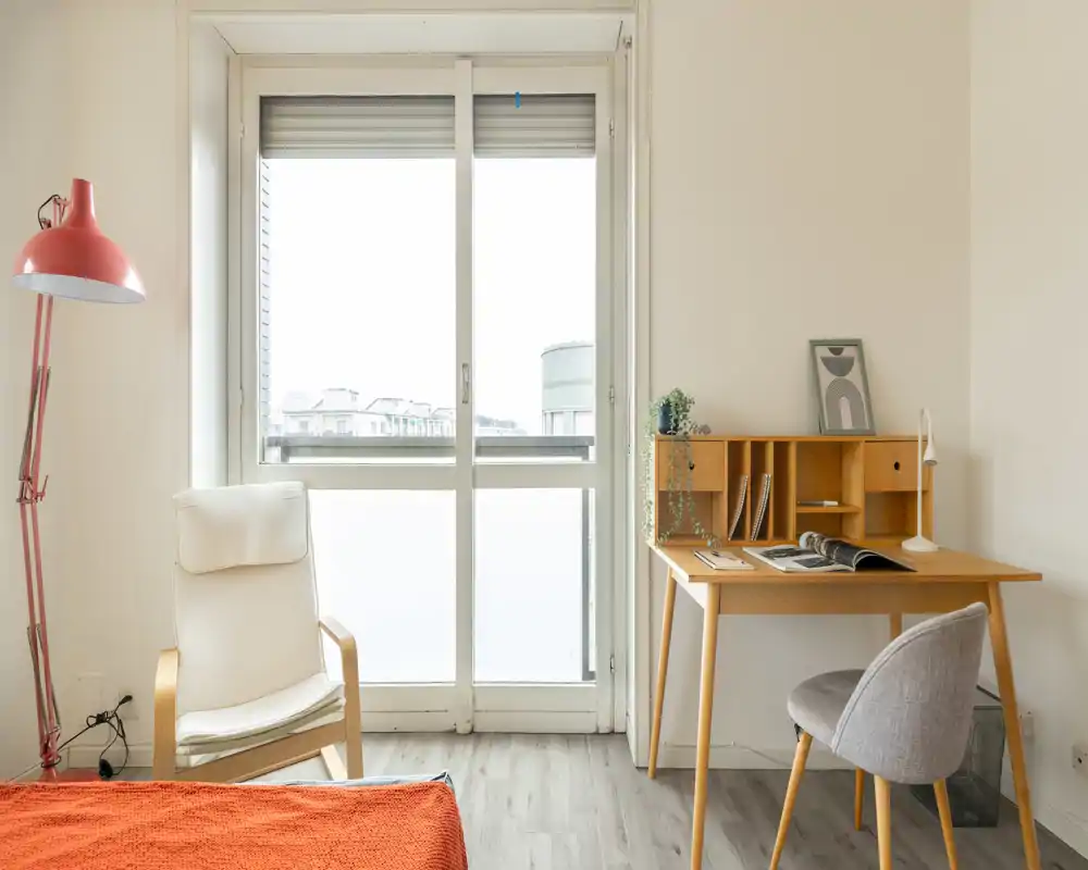 Bright, minimalist bedroom corner with a comfortable armchair, visible bed edge with orange throw, and a tidy wooden desk by a large window letting in abundant natural light.
