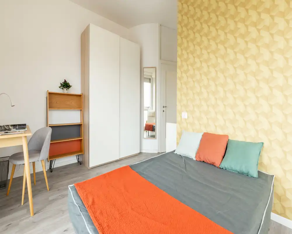 Bright, modern bedroom with a double bed dressed in grey and warm orange textiles, decorative cushions and a patterned yellow accent wall. A wardrobe, desk and chair complete the neat, well-lit space — ideal for rental listings highlighting comfort and contemporary styling.