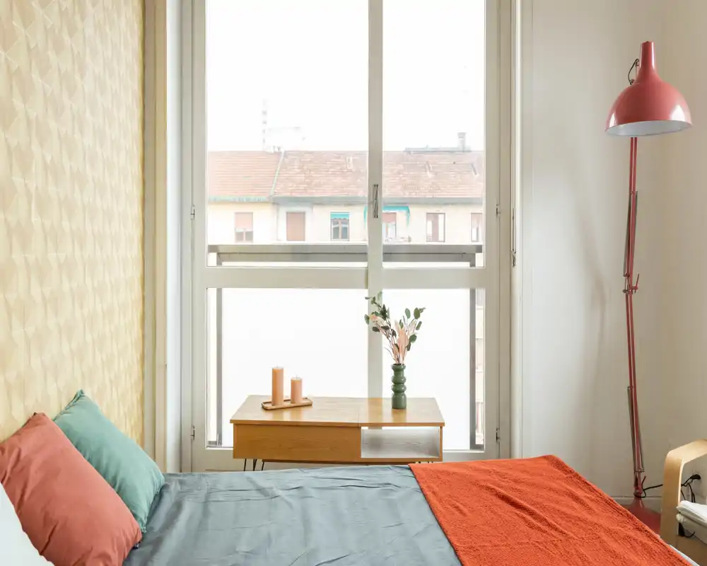 Bright, styled bedroom with a large window, mid-century nightstand, decorative vase and warm textiles including pillows and an orange throw.