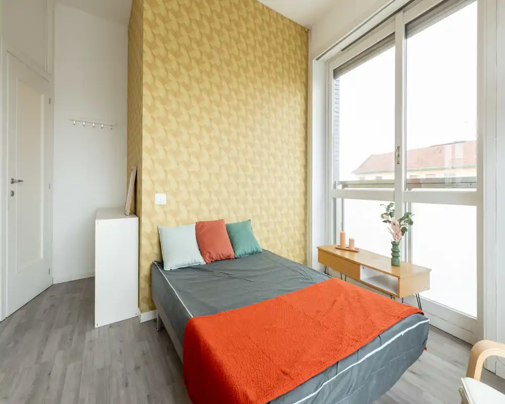 Bright, modern bedroom with a double bed dressed in dark linens, colorful throw and cushions, a patterned feature wall and large window providing abundant natural light.