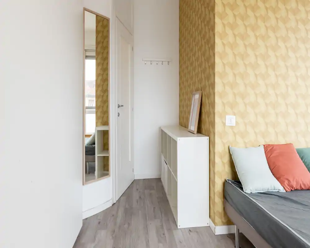 Bright, stylish small bedroom corner with patterned accent wall, a visible mattress with cushions and tidy storage shelving — good natural light.