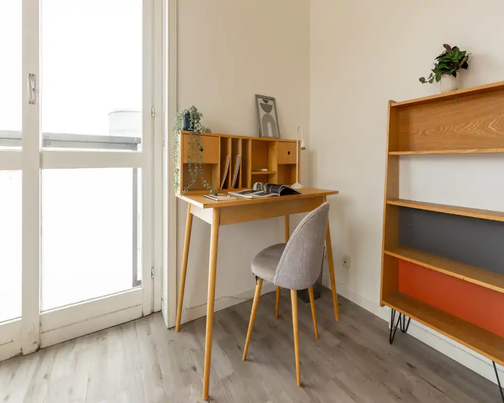 Bright, minimalist living area with a wooden desk and cozy upholstered chair by a large glass door — ideal as a home office corner.