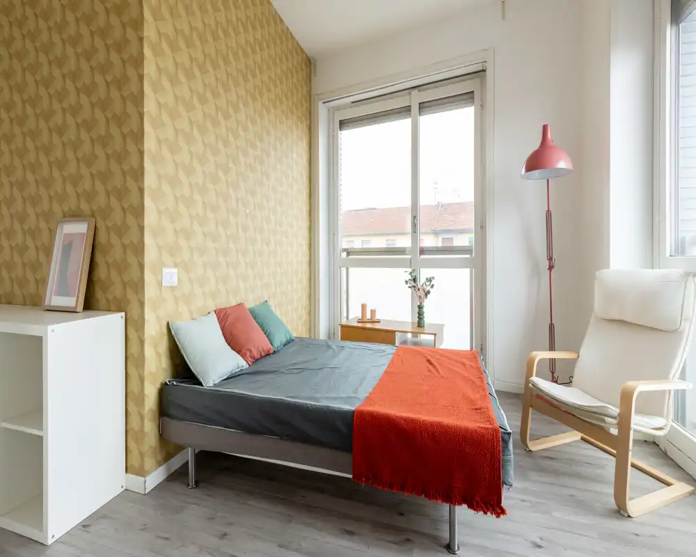 Bright, modern bedroom with a double bed dressed in grey linens and a red throw, large window providing natural light, cozy armchair and minimalist décor.