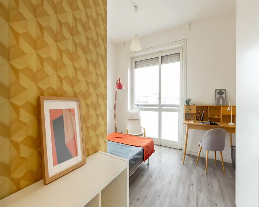 Bright, modern single bedroom with a patterned accent wall, a neatly made single bed and a small desk by the window — ideal for a student or solo tenant.