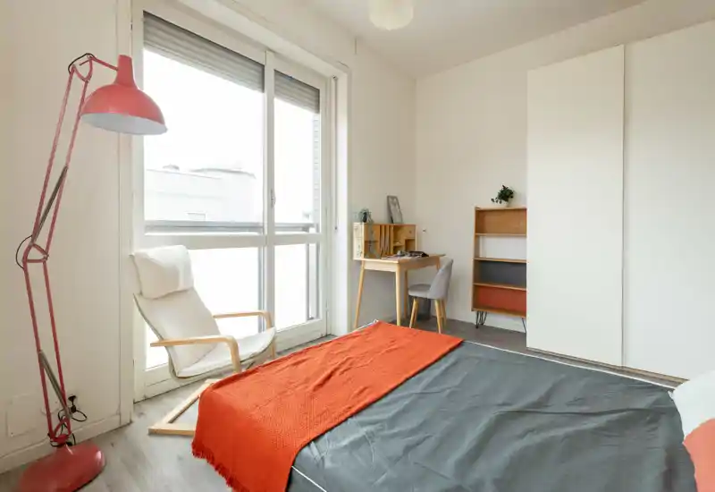 Bright, modern bedroom with a comfortable double bed dressed in grey and an orange throw, a reading chair by a large window and a compact desk with shelving.