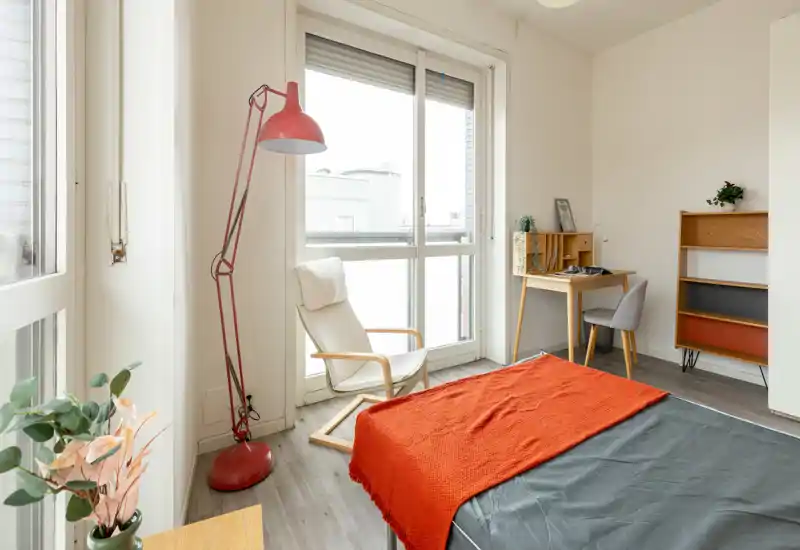 Bright, modern bedroom with large windows, a comfortable bed dressed with a warm orange throw, a reading chair and a small desk — ideal for a cozy single or couple.