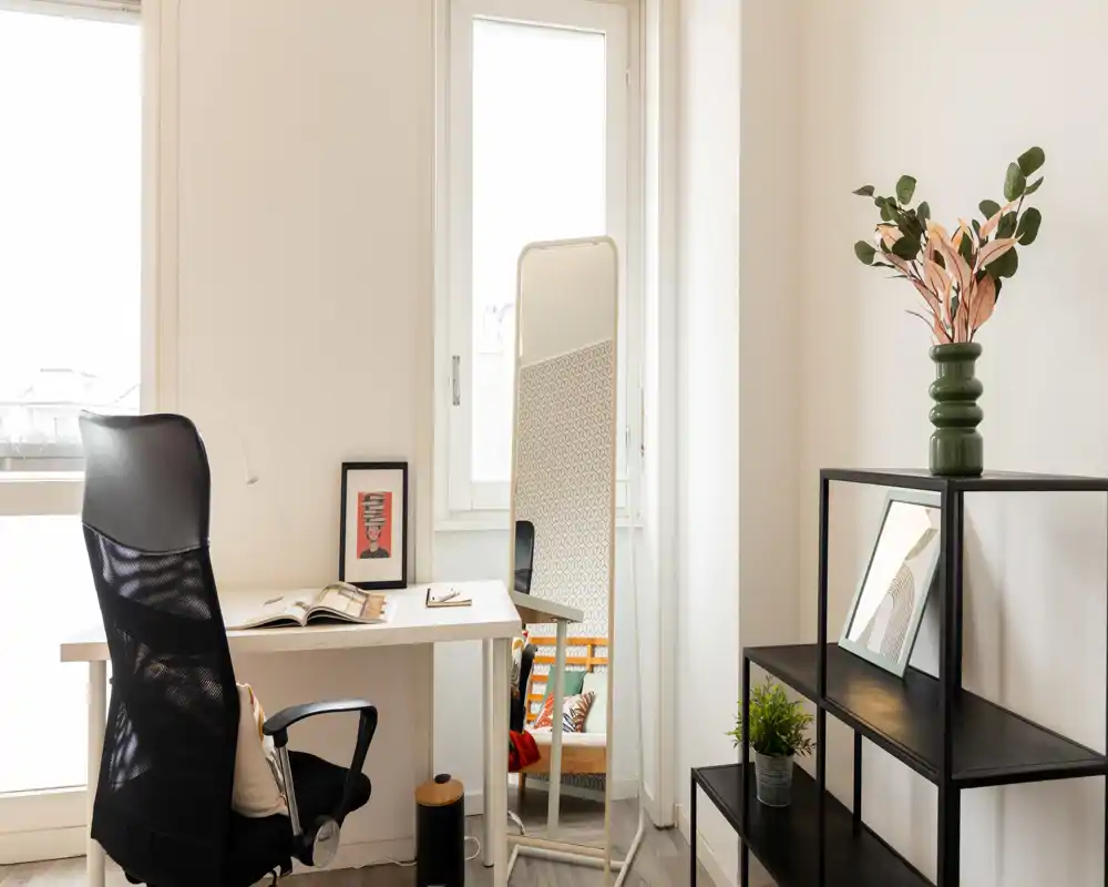 Bright, minimal bedroom corner with a desk and full-length mirror that reflects part of the bedding area; simple shelving and plants add a fresh, functional atmosphere.