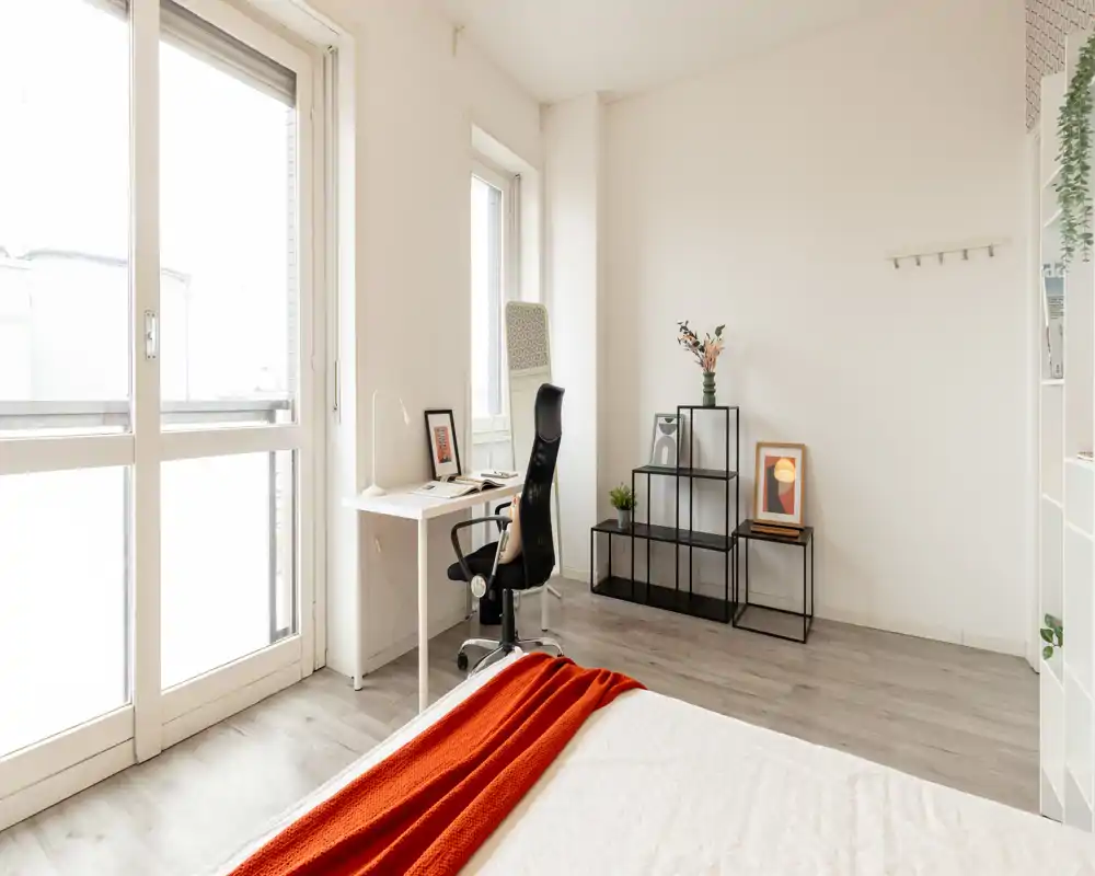 Bright, minimalist bedroom with a large window, a neatly made bed with a warm orange throw, and a compact workspace by the window — ideal for light-filled listings.