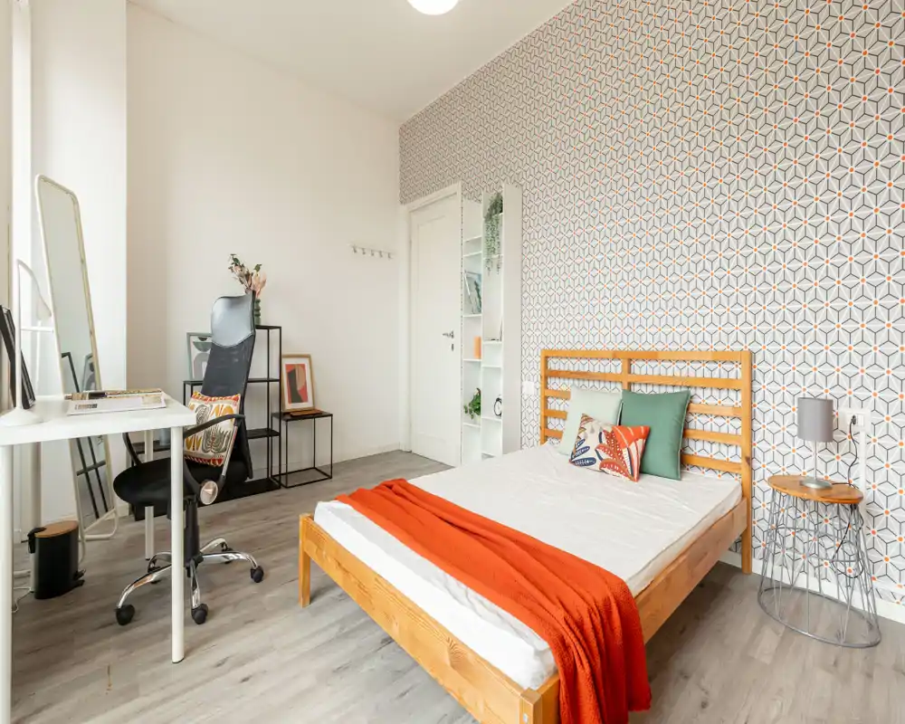 Bright, modern bedroom with a wooden double bed, colorful pillows and an orange throw, plus a small desk and chair by the window. Patterned feature wall and light wood flooring create a fresh, welcoming atmosphere ideal for rentals.
