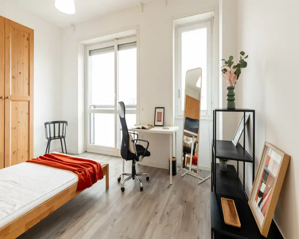 Bright, minimalist bedroom with a wooden single bed, desk by large windows and a cozy red throw.