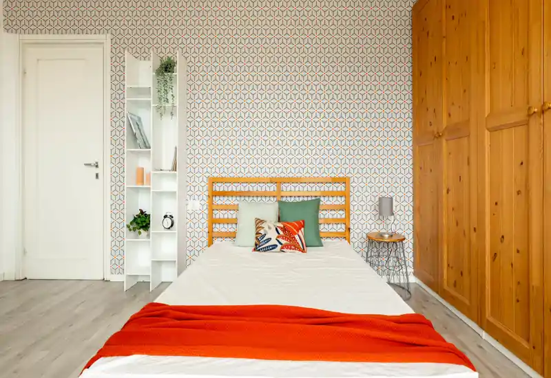 Bright, styled bedroom with a wooden headboard, colorful cushions and an orange throw. The room features patterned wallpaper and a large wooden wardrobe, creating a warm, welcoming atmosphere.