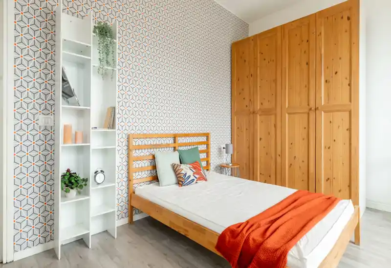 Bright, stylish bedroom with a wooden bed frame, patterned accent wall and a large wardrobe. The room features colorful cushions and a warm orange throw for a welcoming atmosphere.