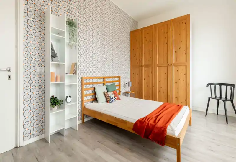 Bright, neatly styled bedroom with a wooden bedframe, colorful cushions and a warm orange throw; large wardrobe and patterned accent wall add character.