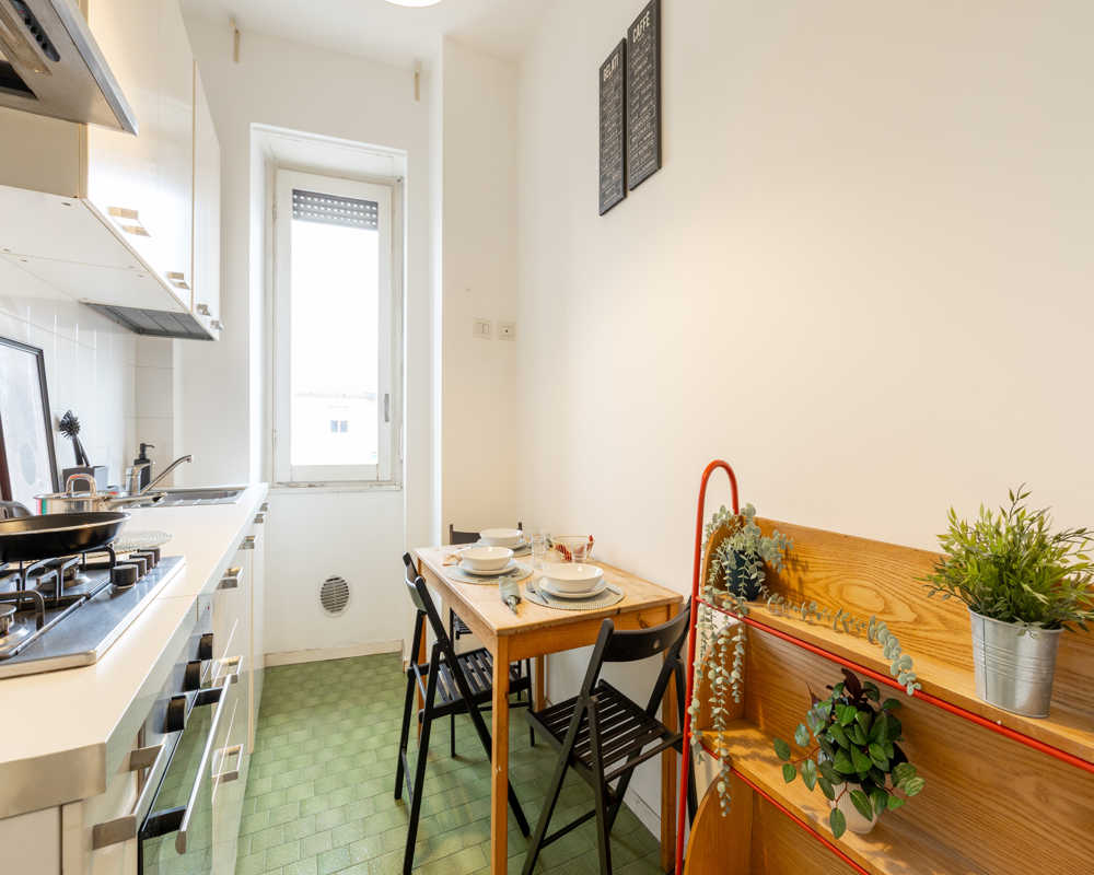 Bright, compact kitchen with built-in stovetop, sink and a small dining table set for two; decorative plants and wooden shelving add warmth.