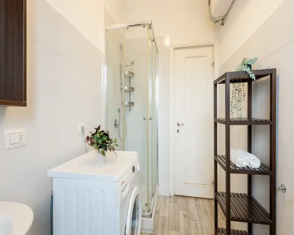 Compact, modern bathroom with a glass shower enclosure, washing machine, and wooden shelving for towels and toiletries. Bright, neutral tiles and a clean layout make the space functional and easy to maintain.