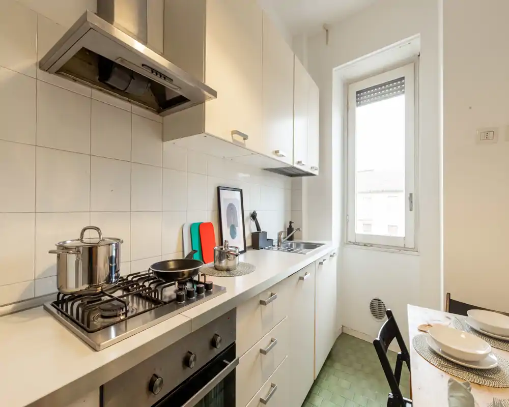 Compact, modern kitchen with a gas hob, stainless hood, sink and a small dining area next to a bright window — practical layout for cooking and casual meals.