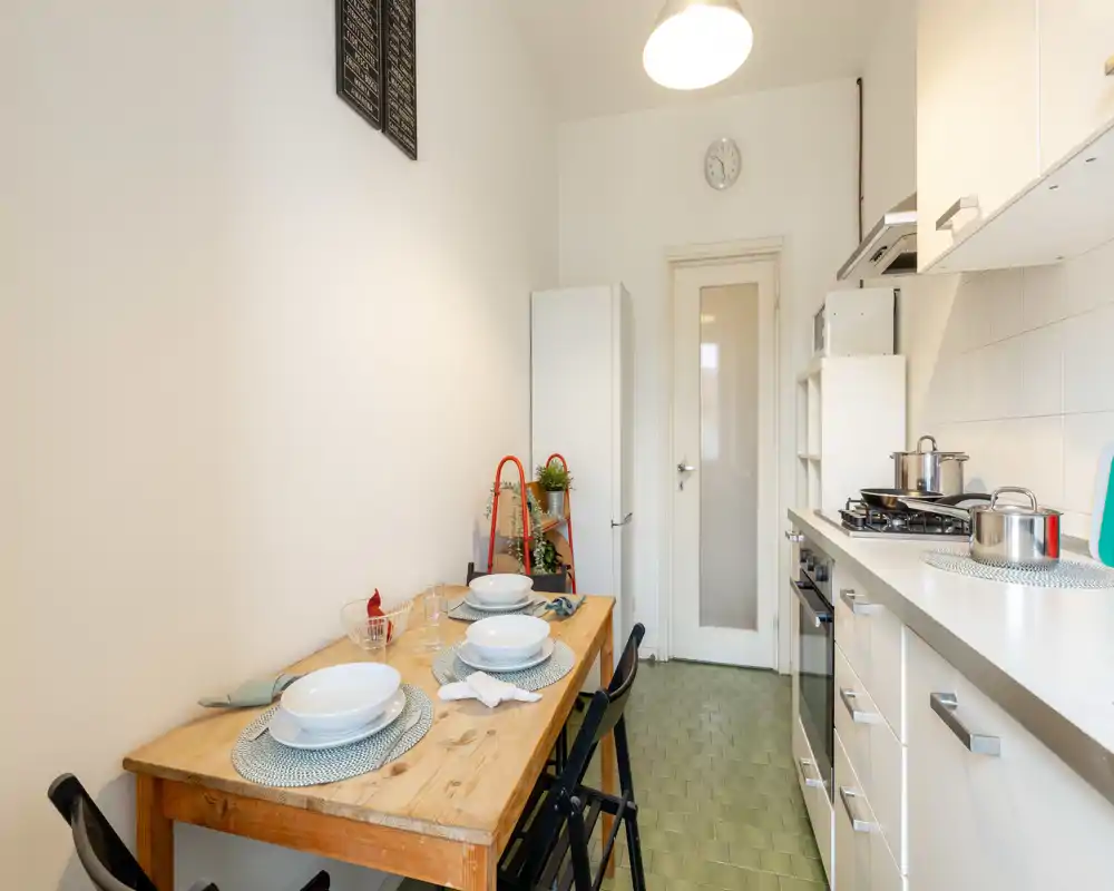 Bright, compact kitchen with a wooden dining table set for four, white cabinetry, and a gas stove. The layout is efficient and well lit, ideal for small apartments.