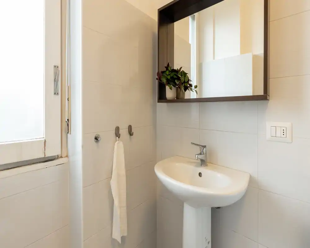 Bright, modern small bathroom with a pedestal sink, wall-mounted mirror with shelf and a towel hanging beside a frosted window.