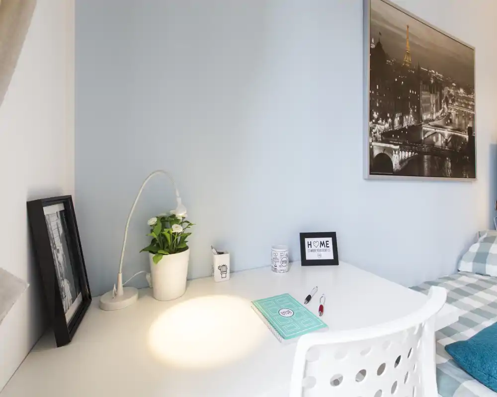 Angolo camera da letto luminoso e accogliente con scrivania bianca, lampada e letto in ordine con copriletto a quadri — perfetto per lavorare e riposare.