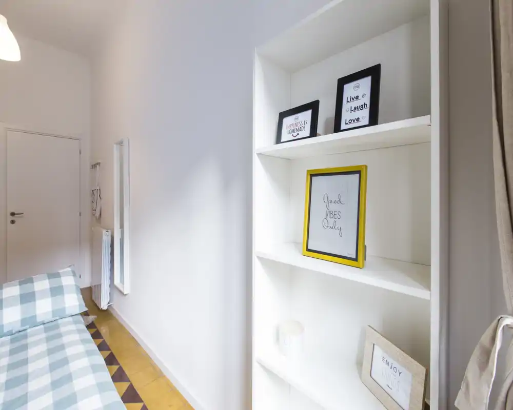 Angolo camera da letto luminoso e minimale con copriletto a quadri e scaffale bianco con cornici: ambiente pulito, moderno e adatto a soggiorni brevi.