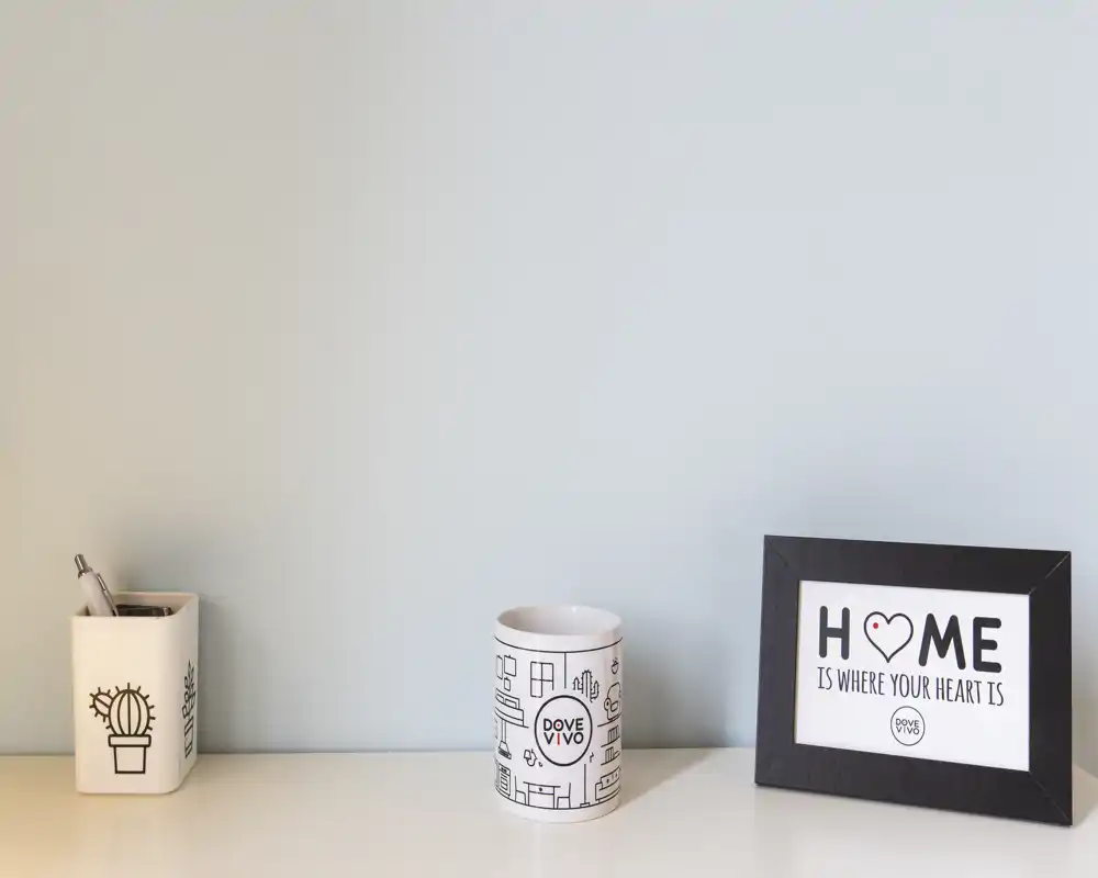 Piccolo angolo decorativo con porta penne, tazza marchiata e cornice con la scritta "Home is where your heart is" su parete chiara — un elemento curato ideale per descrivere un piccolo spazio di lavoro o l’ingresso.