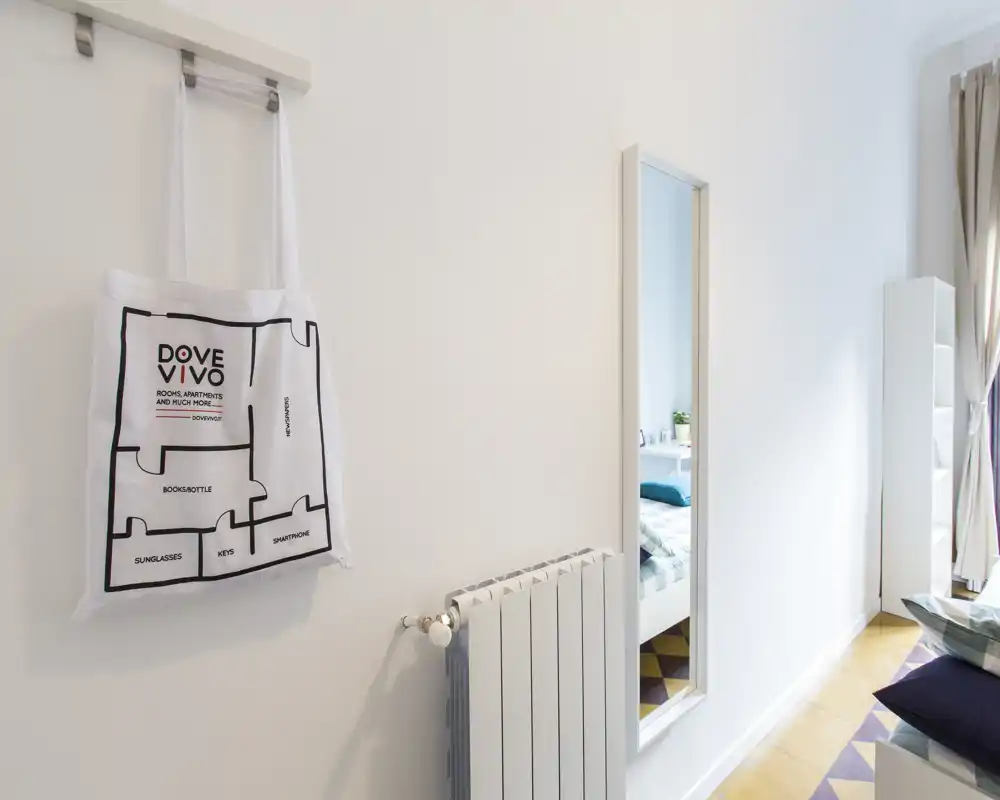 Angolo camera luminoso e minimale con bordo del letto visibile, specchio a figura intera e pavimento in legno chiaro; arioso e moderno.