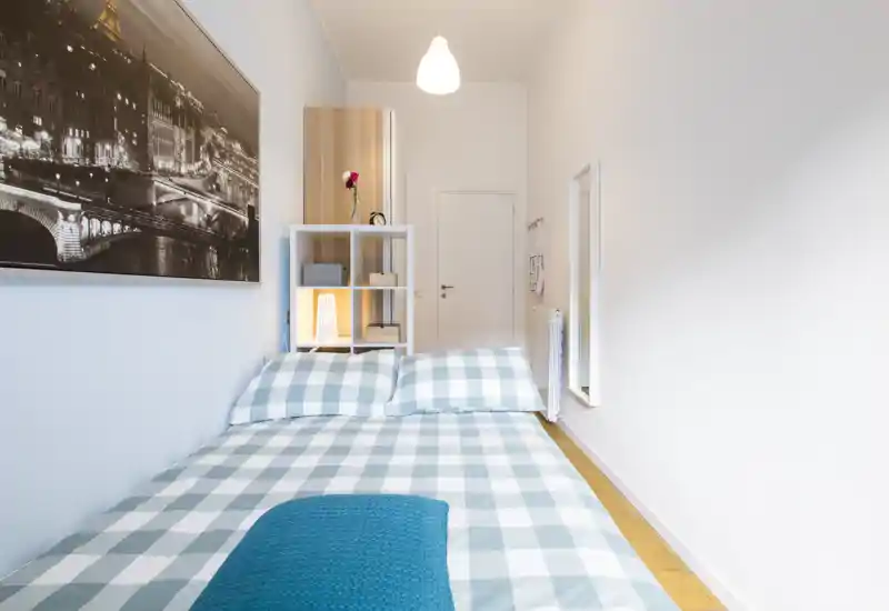 Camera da letto luminosa e ben allestita con letto matrimoniale con lenzuola a quadri, mensola con lampada e decorazioni e una grande foto incorniciata sulla parete — perfetta per un annuncio accogliente.