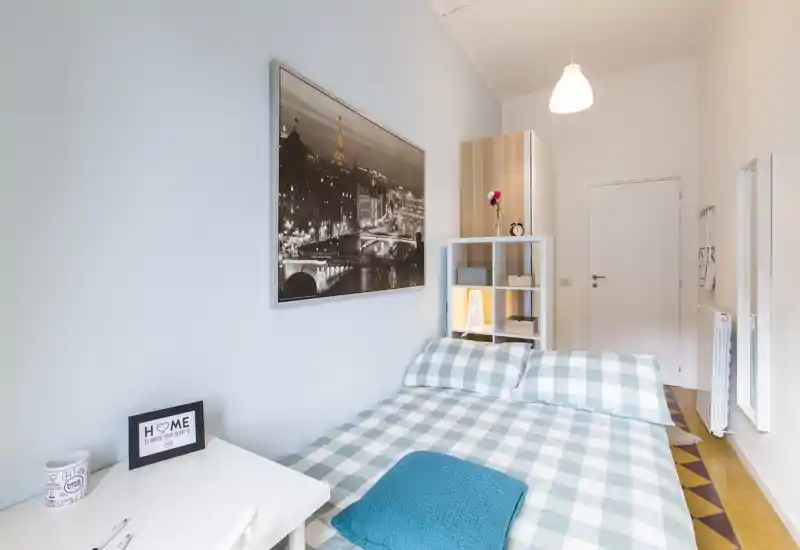 Camera da letto luminosa e ben arredata con letto matrimoniale in copriletto a quadri, scaffale portaoggetti con lampada soffusa e grande quadro sopra la testiera.
