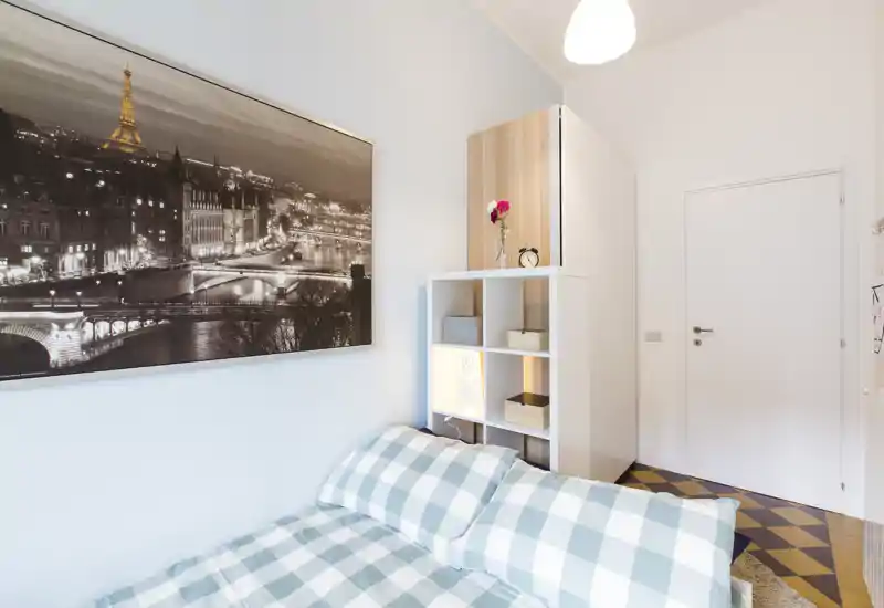 Camera da letto compatta e luminosa con copriletto a quadri, mensole decorative e un grande quadro in bianco e nero di Parigi sopra il letto — accogliente e ben illuminata per soggiorni brevi.