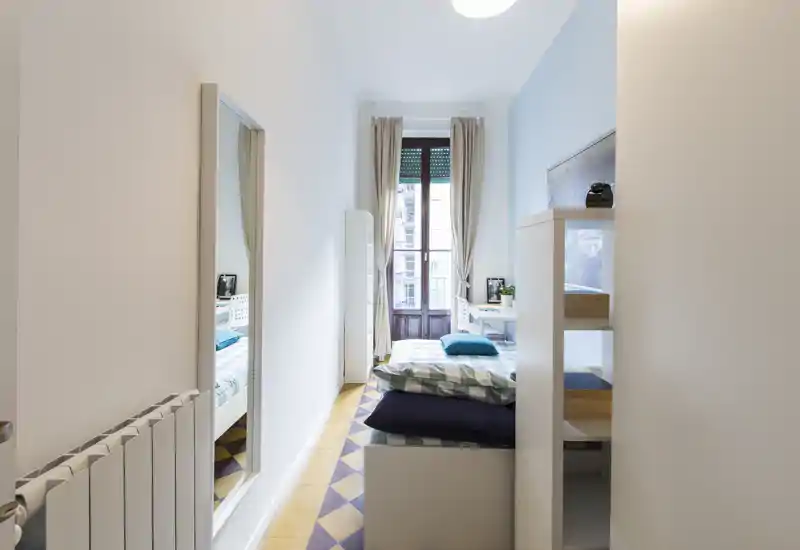 Camera da letto luminosa e stretta con letto singolo rivolto verso una finestra grande. Arredo minimalista, tessili accoglienti e luce naturale creano un’atmosfera invitante ideale per un ospite singolo.