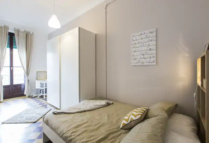 Camera da letto luminosa e curata con letto matrimoniale ben rifatto, cuscini decorativi e armadio. La luce naturale dalla porta del balcone e l’illuminazione interna creano un’atmosfera calda e accogliente.