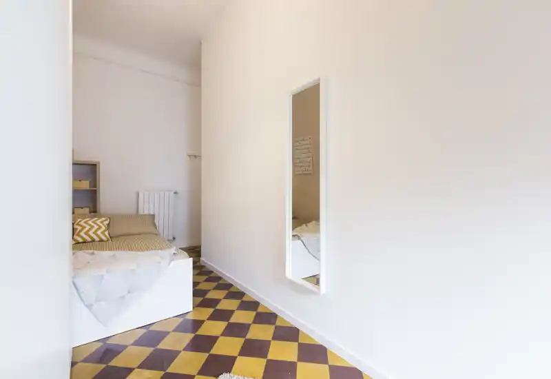 Angolo camera da letto luminoso e minimale con un letto singolo in una nicchia, pavimento a scacchiera e uno specchio a figura intera che riflette parte del letto.
