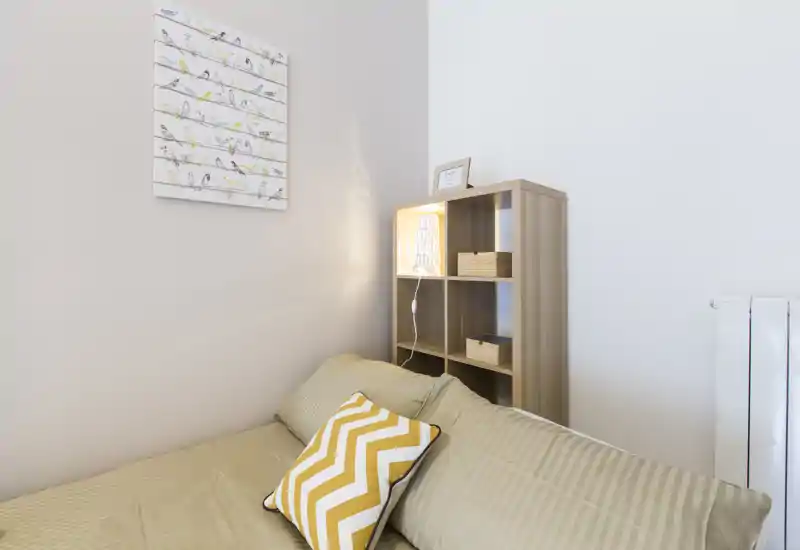 Angolo camera compatto e ben illuminato con letto rifatto, cuscino decorativo e piccolo scaffale aperto con lampada — luminoso, moderno e accogliente.