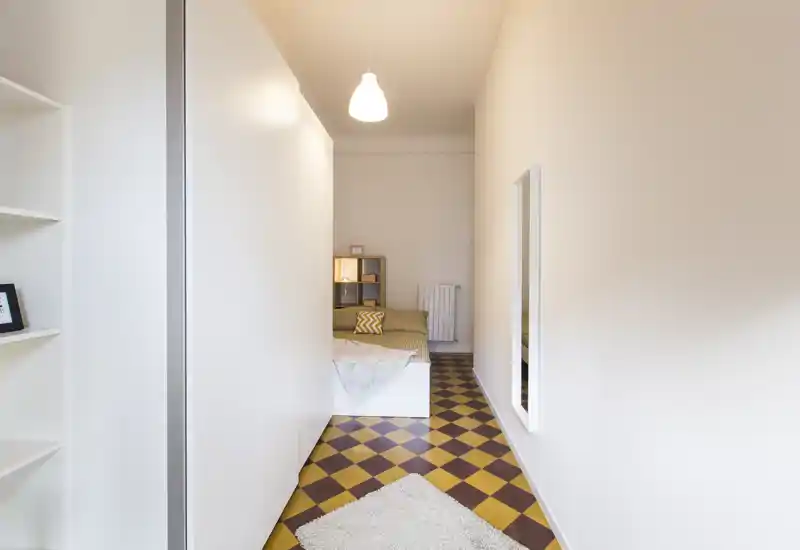 Camera da letto compatta e luminosa con letto singolo, pavimento a piastrelle a scacchiera e arredi minimal; una mensola e una luce soffusa rendono l’ambiente accogliente.