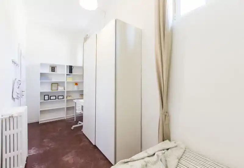 Angolo di camera compatto con un letto singolo parzialmente visibile in primo piano, un armadio alto e scaffalature con area scrivania sullo sfondo. Luminoso, stile minimale con toni neutri e una postazione funzionale.