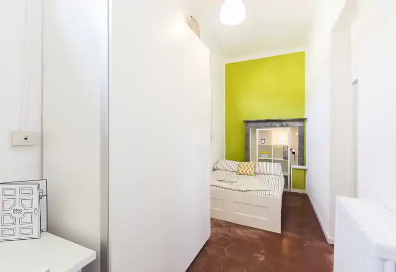 Camera compatta con letto singolo ben rifatto, biancheria a righe e una vivace parete verde che crea un'atmosfera allegra. La piccola mensola integrata sopra la testiera e l'illuminazione soffusa rendono lo spazio pratico e accogliente.