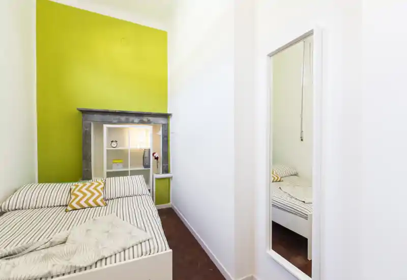 Piccola camera da letto luminosa con letto a righe, parete d'accento verde e mensola decorativa incorniciata da un finto caminetto. Biancheria pulita e uno specchio grande ampliano lo spazio compatto.