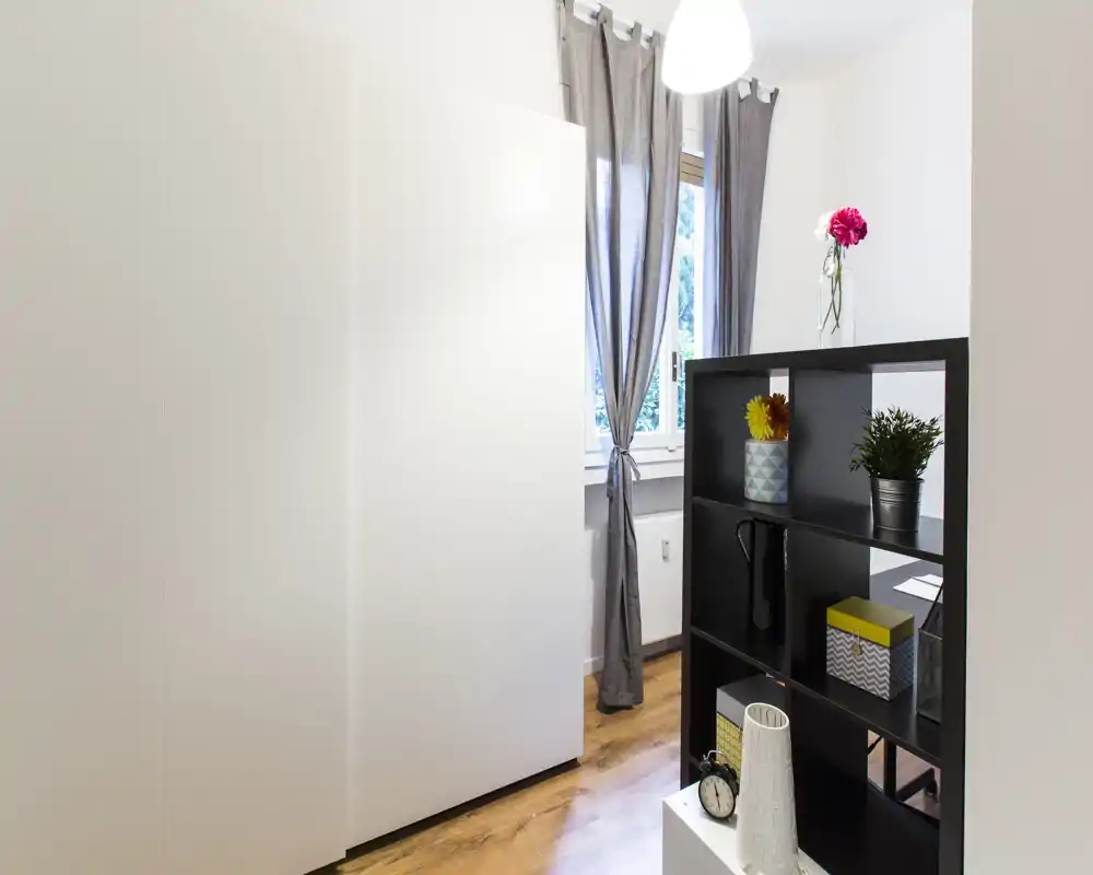 Zona giorno luminosa e ordinata con una libreria divisoria, pavimento in legno e una finestra con tende che illumina l'ambiente.