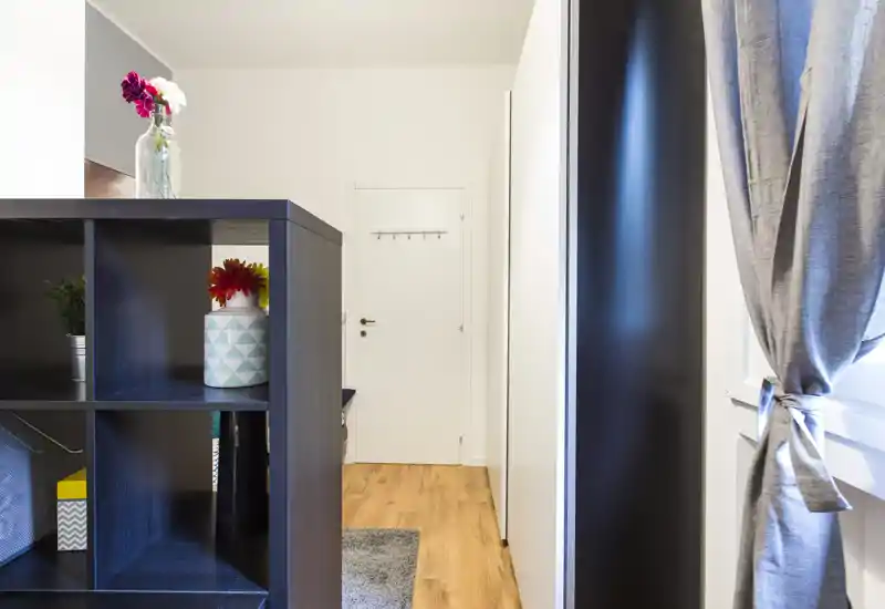 Neat entrance corridor with a freestanding shelf divider, light wood floor and tied-back curtains providing a tidy, welcoming entry.