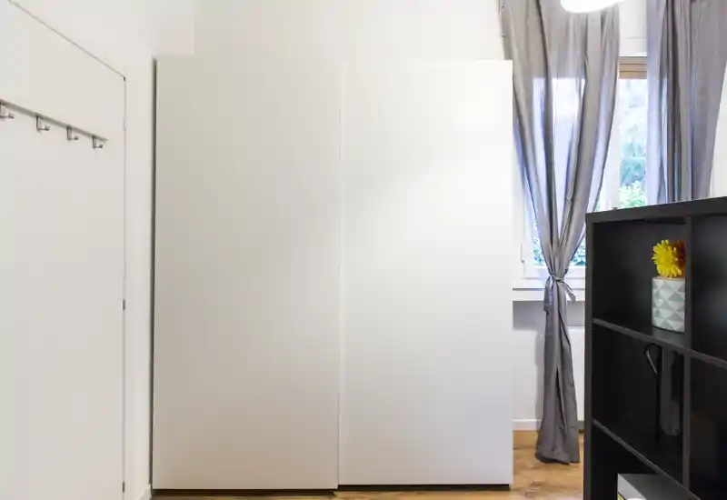 Angolo di camera minimal con grandi armadi bianchi, una finestra con tende grigie e una piccola libreria con oggetti decorativi; luminoso, pulito e moderno.