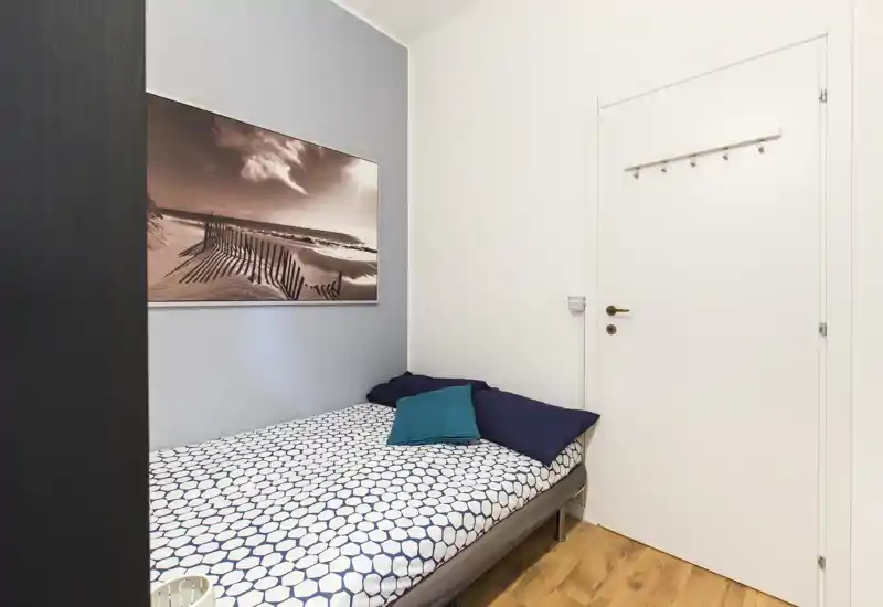 Camera singola compatta con letto a una piazza dal copriletto a motivi, cuscini turchese e blu scuro, quadro con paesaggio marino e pavimento in legno caldo; luminosa e arredata in modo semplice.
