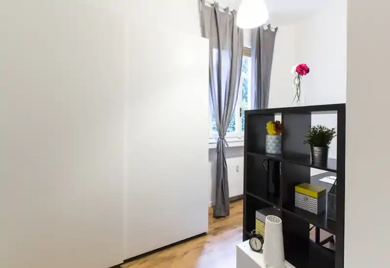 Bright, tidy living area with a freestanding shelving unit dividing the space, wooden floors and a curtained window that brings in natural light.