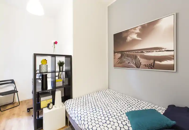 Camera da letto luminosa e moderna con letto matrimoniale, copriletto a motivo e cuscini decorativi; scaffalatura aperta e quadro costiero completano l'ambiente.
