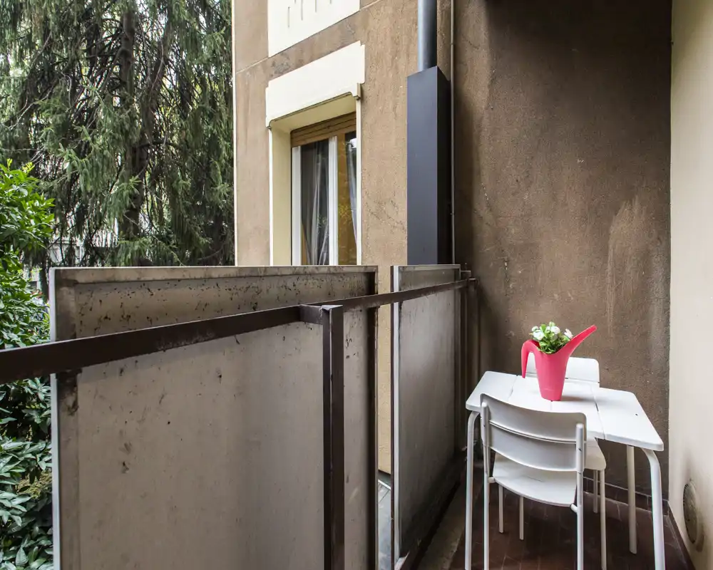 Piccolo balcone privato con un tavolino e due sedie compatte, decorato con un annaffiatoio rosa con pianta; vista sul verde e sull'edificio vicino.