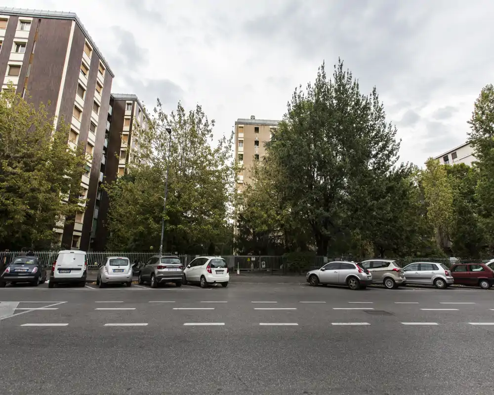 Vista esterna verso la strada con auto parcheggiate, alberi ed edifici residenziali — utile per mostrare la facciata di quartiere e la disponibilità di parcheggio.