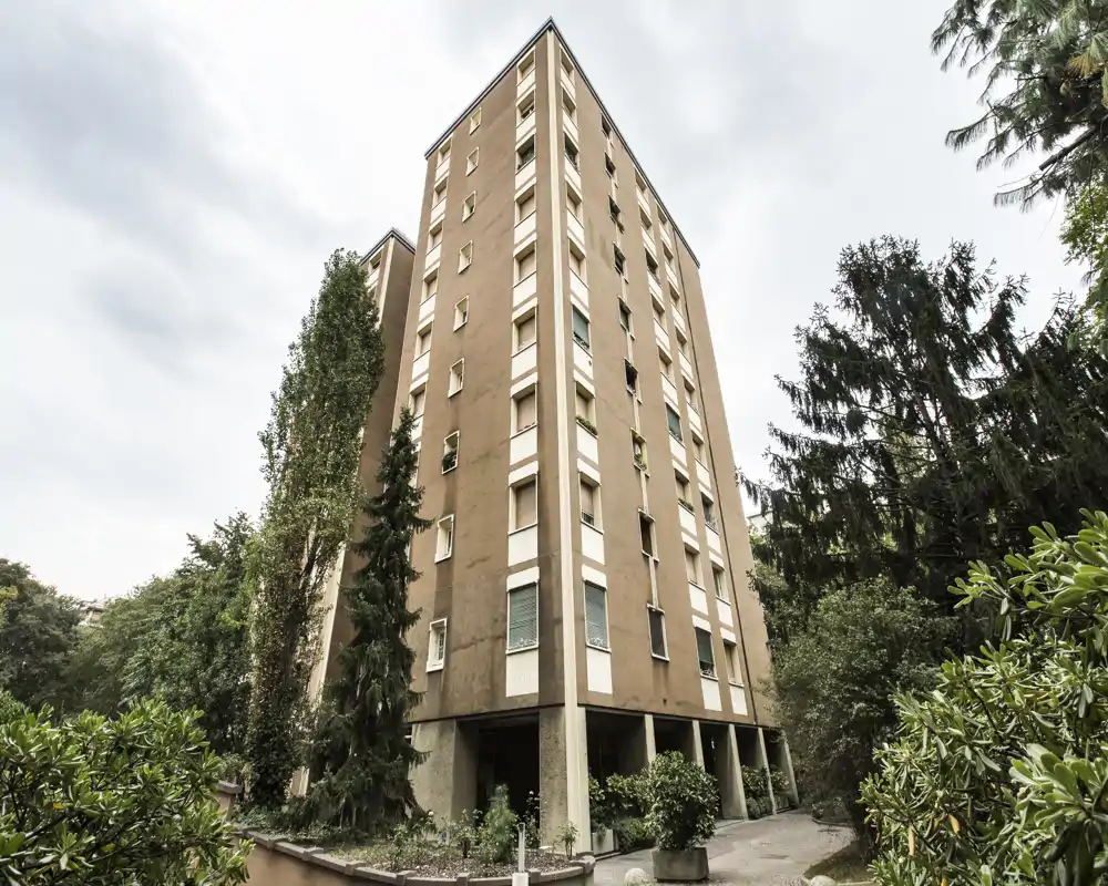 Vista esterna di un edificio residenziale di media altezza circondato da alberi maturi e giardini; la foto mette in evidenza la facciata e l’area d’ingresso con luce soffusa.