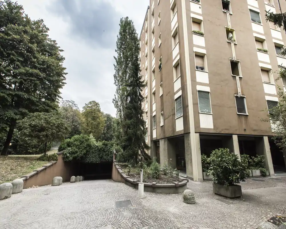 Cortile esterno e rampa d'accesso a un garage di un edificio residenziale, con alberi e aree verdi circostanti.
